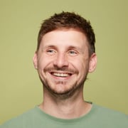 Porträt eines lächelnden Mannes mit kurzen braunen Haaren, Bart und Ohrsteckern, vor einem hellgrünen Hintergrund. Er trägt ein grünes T-Shirt und schaut fröhlich in die Ferne