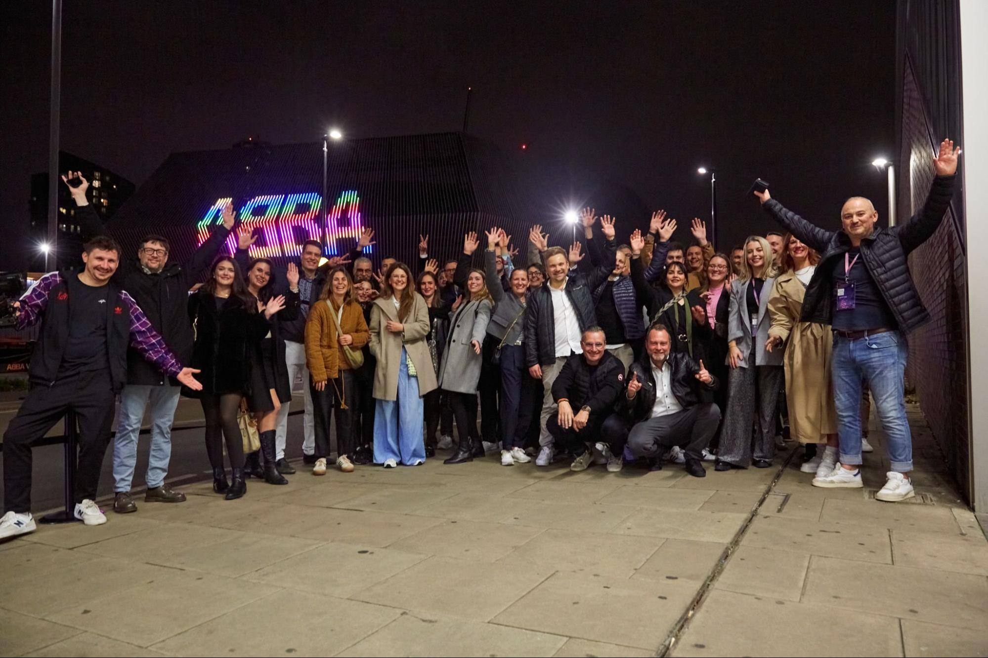 ONEDOT Gruppenfoto: Viele fröhliche Menschen schauen lachend und jubelnd in die Kamera. Im Hintergrund ist die bunte Leuchtschrift vom ABBA-Event zu sehen.